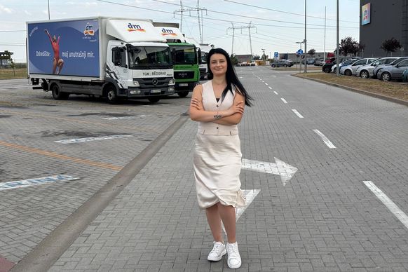 Milica u svetlo beloj haljini stoji na parkingu dm-a, iza nje su dm kamioni